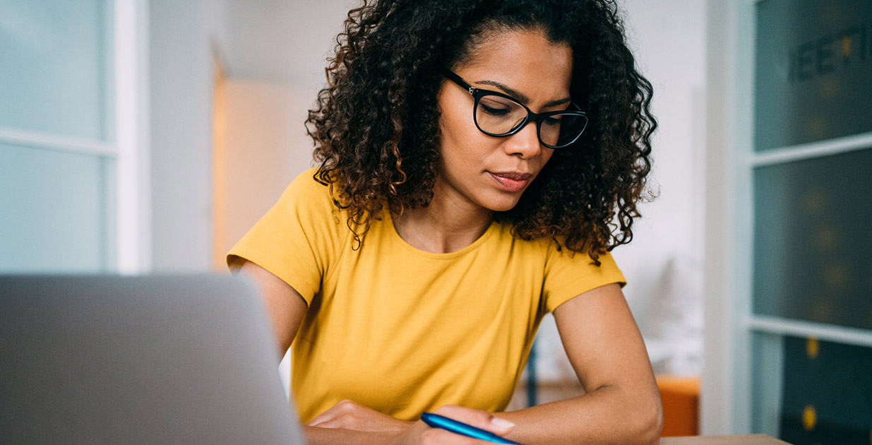 Woman in yellow shirt with glasses sitting in front of computer writing with a pen.