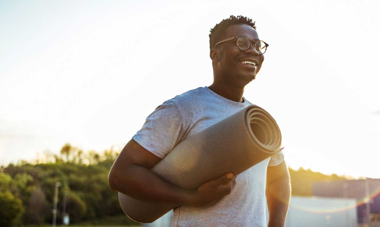 Man wearing glasses smiling holding yoga mat under arm